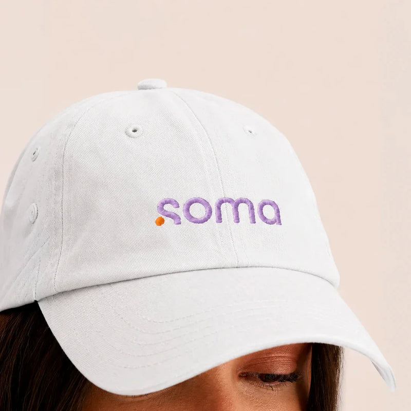 Gorra blanca con logo bordado 'soma' en morado y naranja, estilo casual y moderno