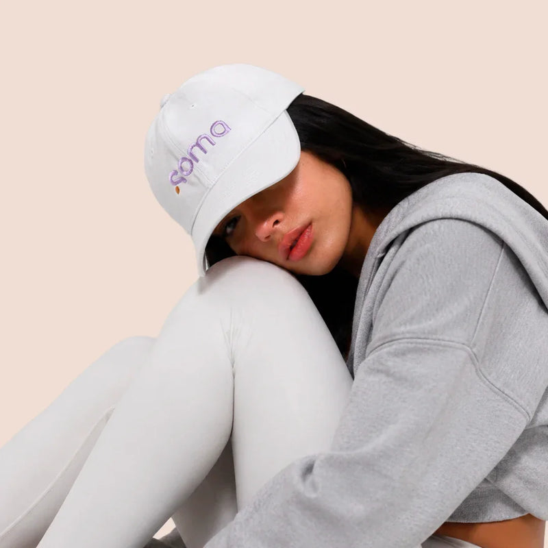 Mujer con gorra blanca bordada 'soma', sudadera gris y leggings blancos, look casual cómodo