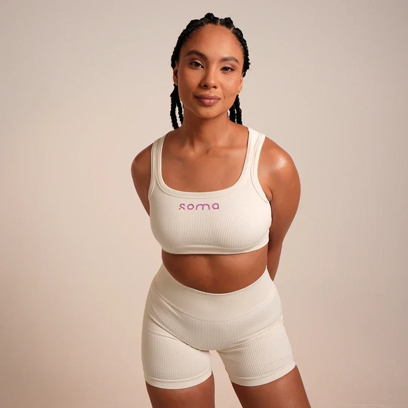 Mujer con conjunto deportivo blanco ribeteado, top con logo Soma y shorts, ideal para yoga o entrenamiento.